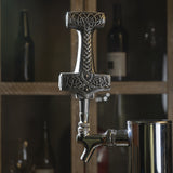 A close-up of a metal beer tap handle shaped like a Viking sword with ornate Celtic knotwork engravings, mounted on a stainless steel tap fixture in a bar setting with blurred bottles in the background.