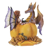 A whimsical fantasy figurine of a fairy with purple wings sitting on a large orange pumpkin, facing a purple and gold dragon with black-and-white striped horns, set against a plain white background.