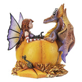 A whimsical fantasy figurine featuring a fairy with purple wings and red hair sitting on a large yellow pumpkin, facing a purple and gold dragon with black-and-white striped horns. A small black cat rests on the pumpkin, and a textured base with leaf-like details surrounds the scene.