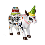 A decorative ceramic cow figurine painted white with colorful polka dots, black markings, a green party hat, and a birthday cake on its back. The cow is posed on a plain white background.