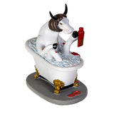 A decorative figurine of a white cow with brown and black horns sitting in a white bathtub filled with blue bubbles, holding a red soap bottle, on a gray base with gold feet.