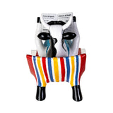 A novelty ceramic cow-shaped magazine holder and newspaper rack, featuring a black and white cow body, colorful red, yellow, blue, and white striped seat, and black hooves, holding two mock newspapers against a white background.