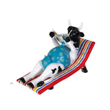 A decorative figurine of a black-and-white cow lounging on a striped beach chair and reading a newspaper, shown on a plain white background.