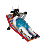 A decorative figurine of a black-and-white cow lounging on a red chaise lounge with blue, yellow, and white stripes, reading a newspaper. The cow is wearing a blue polka-dot swimsuit.