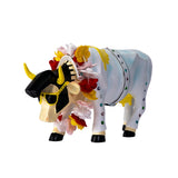 A decorative cow figurine or sculpture in white with black and yellow details, decorated with red, yellow, and pink flower garlands, set against a plain white background.