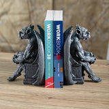 A pair of dark gray gothic dragon bookends holding up two blue and teal books on a wooden tabletop, with a softly blurred neutral background.