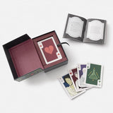 A boxed card game set displayed on a light gray background, including an open dark box with a card deck inside, an open booklet, and several illustrated cards fanned out in front.