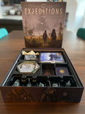 Board game box titled 'Expeditions' with game pieces and cards on a wooden table.