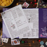 Game components including a purple book with printed pages, cards, and dice on a wooden surface.