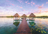 Wooden dock leading to three thatched-roof structures over water with a colorful sky.
