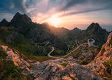 Winding mountain road at sunset with rocky peaks and a dramatic sky.