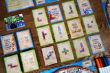A close-up overhead view of a tabletop game setup with multiple illustrated cards arranged on a wooden surface. The cards feature colorful fantasy artwork and text, with some cards showing blue borders and others showing green borders.