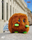 Plush toy resembling a brown creature with large eyes and green feet in front of a building.