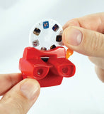 A close-up of a red plastic toy camera with a white circular dial showing small printed sample images, held between two hands against a plain light background.