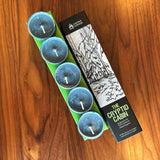Four teal tealight candles in a green plastic tray next to their black and white illustrated packaging box on a wooden table. The box is labeled 'The Cryptid Cabin' by 'Cantrip Candles'.