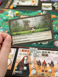 A hand holds a printed game card in front of a board game setup. The card shows a forest scene with a small structure and text rules, while the board game table below includes player tokens, terrain, and other cards.