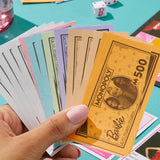Hand holding a Monopoly card featuring Barbie on a colorful table with game pieces.