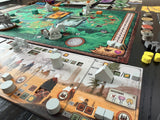 A close-up of a board game setup on a dark tabletop, showing two long game boards with illustrated terrain, white player tokens, stacks of round coins, and yellow dice.