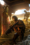 Person in tactical gear holding a weapon in a desert setting with an alien figure and another person in the background.