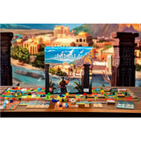 The board game Papyria set up on a wooden table, with the illustrated game box standing upright in the center and colorful game pieces and cards spread out in front. The backdrop is a painted coastal city scene with stone pillars.