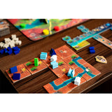 A close-up of a board game set up on a dark wooden table, showing colorful terrain tiles, small buildings, river paths, and player tokens.