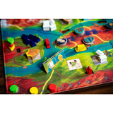 A close-up of a colorful board game board with miniature terrain pieces, player tokens, and cards. The board features a detailed map design with green hills, blue water, and red terrain, and various small toy pieces are scattered across the playing surface.
