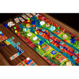 A colorful board game set arranged on a wooden table, with a long illustrated game board, small plastic pieces, tokens, and accessories visible in a close-up overhead view.