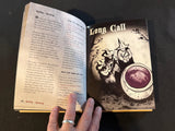 Open book with a 'Long Call' card featuring a witch and a cup on a dark surface.