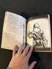 Open book with illustrations of a character and weapon, held by a hand.