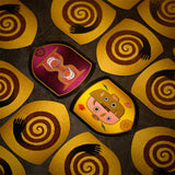 A close-up overhead view of several decorative game pieces or tokens arranged on a dark textured surface. The pieces are irregularly shaped with rounded edges and feature bold illustrated designs: most are yellow with dark brown spiral motifs and black hand-like shapes, while two central pieces show stylized cartoon faces in pink, yellow, brown, and purple tones.