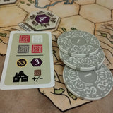 Board game components including a card with symbols and numbers and round tiles on a hexagonal tile background.