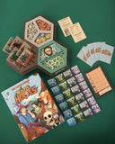 Board game components including cards, a hexagonal board, and a book on a green background