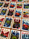 A close-up view of a card game board with multiple illustrated cards arranged in a grid on a brown surface. The cards feature stylized cars, roads, and city scenes in bright colors, with red, blue, green, yellow, and black artwork.
