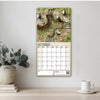 Calendar with nature-themed design on a wall above a table with a plant and cup.